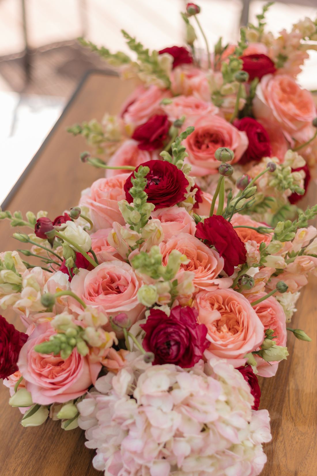A long low centerpiece of garden roses, hydrangea and snapdragons in blush and deep red