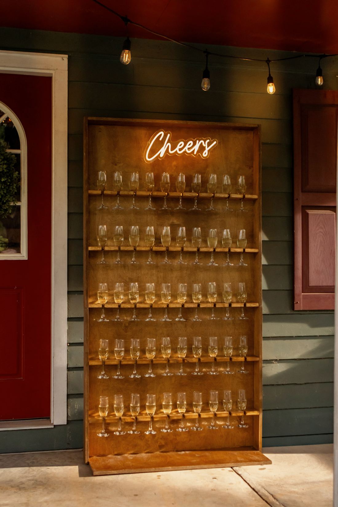A wooden champagne wall of filled flutes beneath a glowing “Cheers” neon sign
