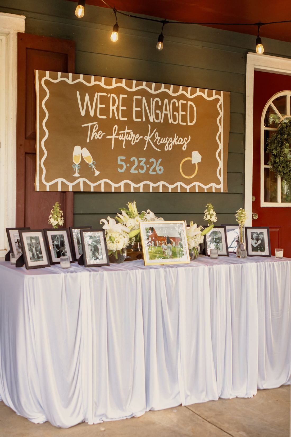 A draped table of framed photographs and white lily arrangements beneath a hand-painted “We’re Engaged” sign