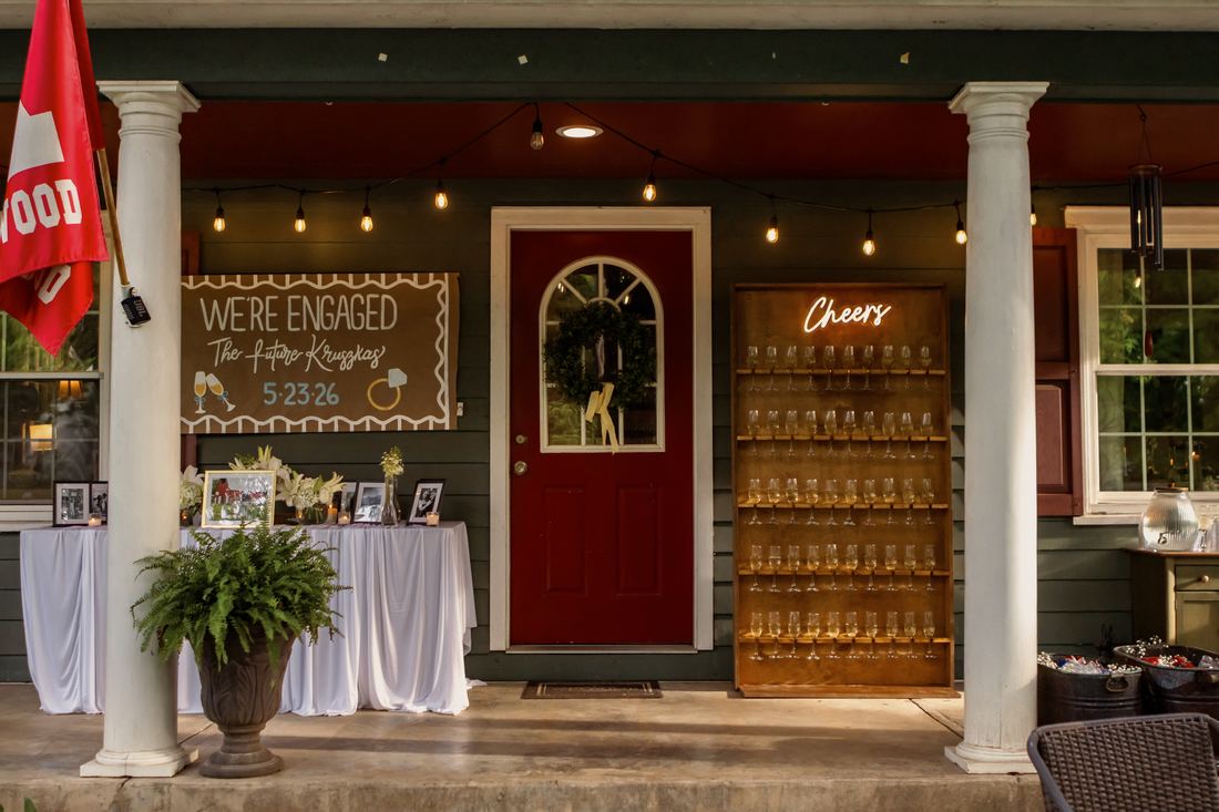 A covered porch set for an engagement party, with a hand-painted sign, a draped table of framed photographs and a champagne wall under string lights