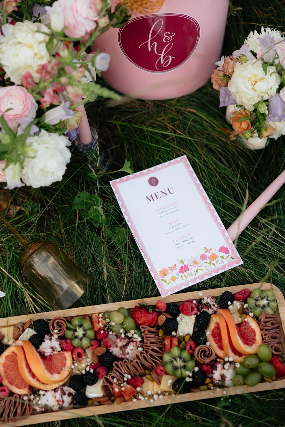 A grazing board laid on grass beside a printed menu, a pink flower tub and white florals