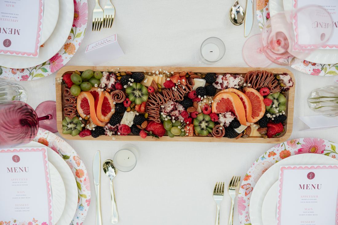 A long grazing board running down a styled table between printed menus and florals