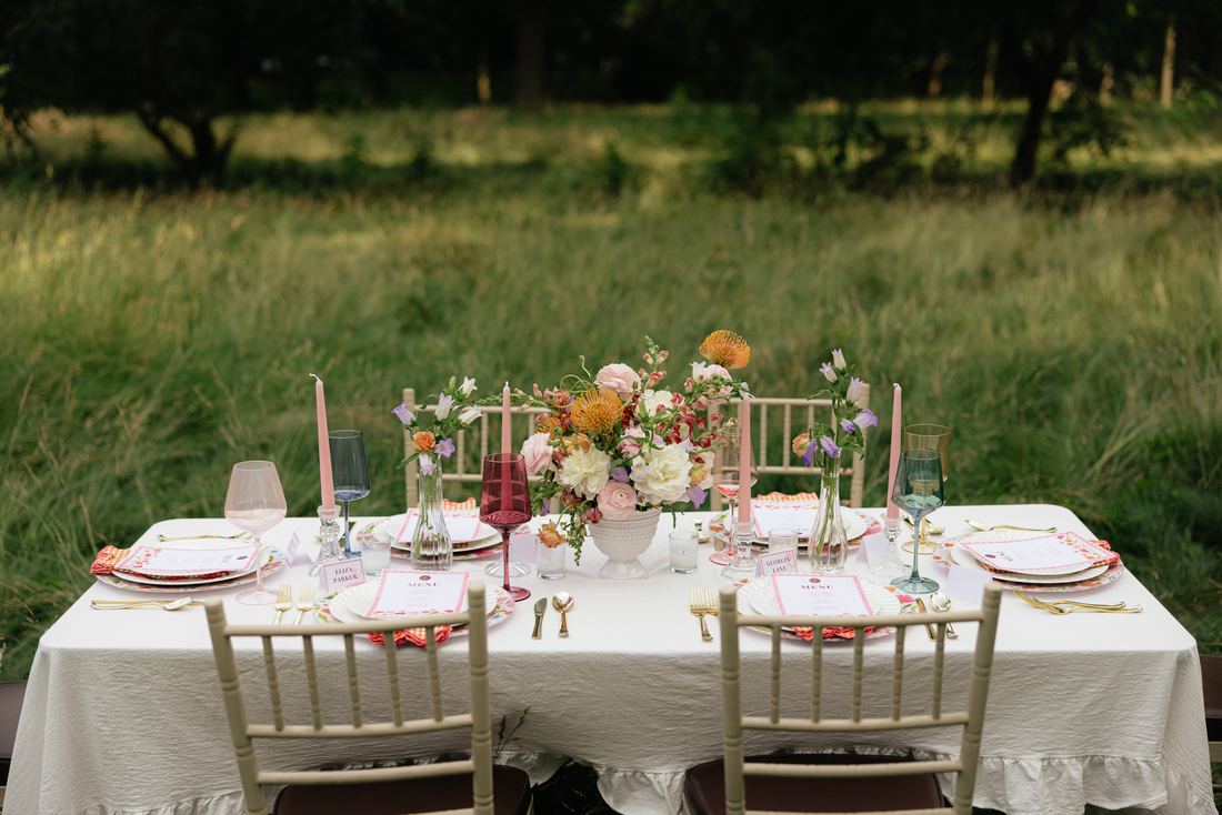 A long white-linen table set with coral florals and gold chairs, standing in a summer meadow