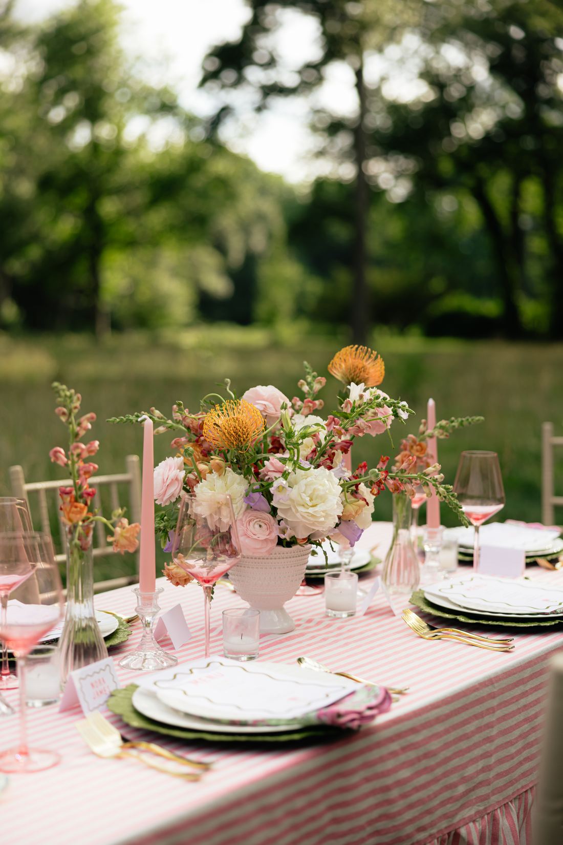 A pink stripe table in a meadow set with a blush centerpiece of garden roses and pincushion protea, pink taper candles and green scalloped chargers