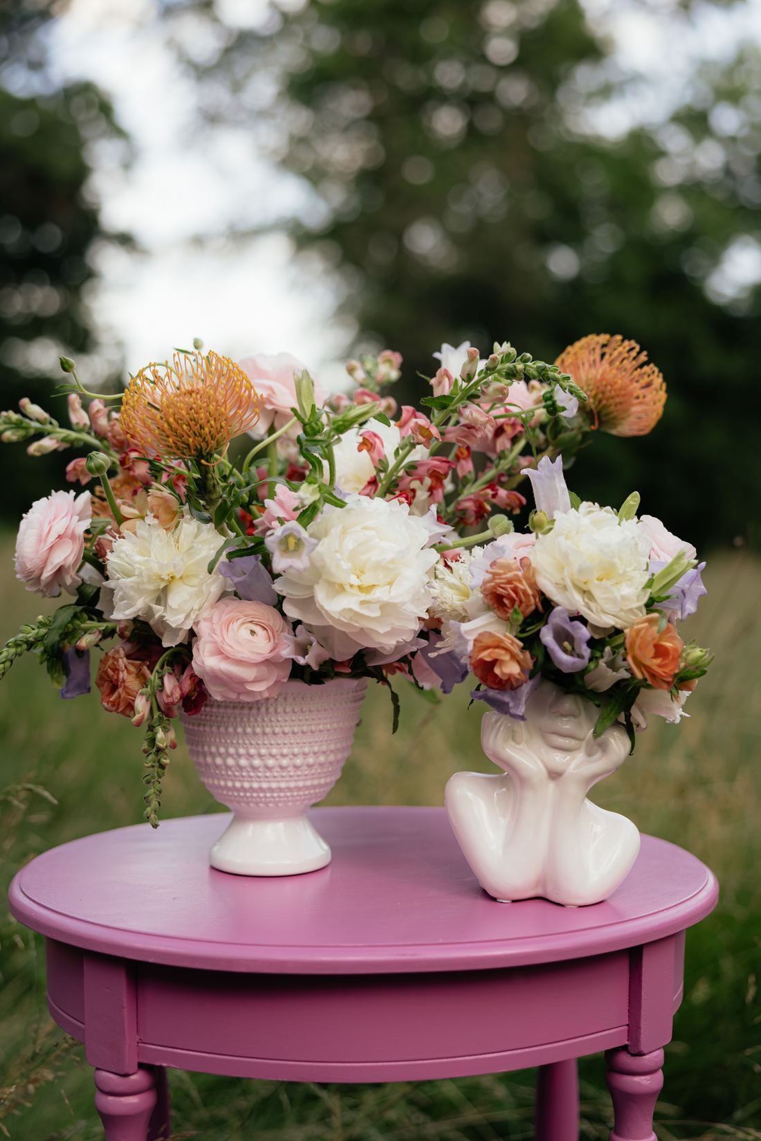 A peach and cream arrangement with pincushion protea in a footed vase on a pink table