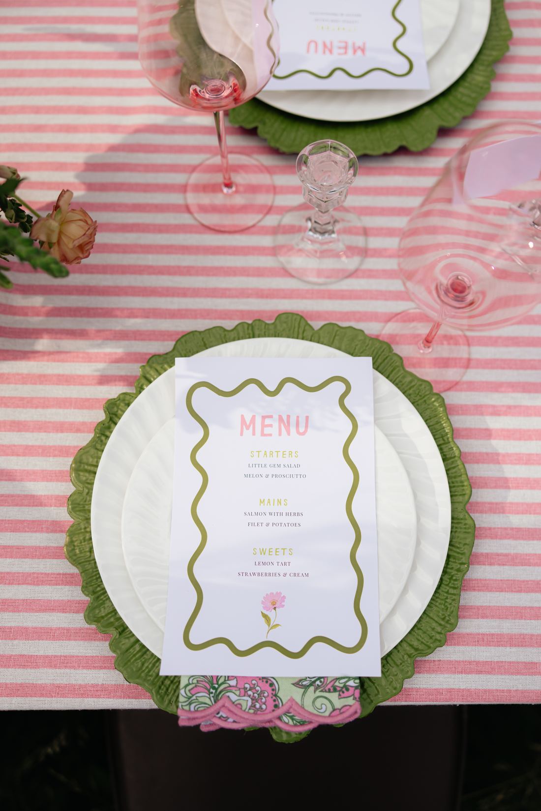 A place setting on pink stripe linen with a green scalloped charger and a printed menu card