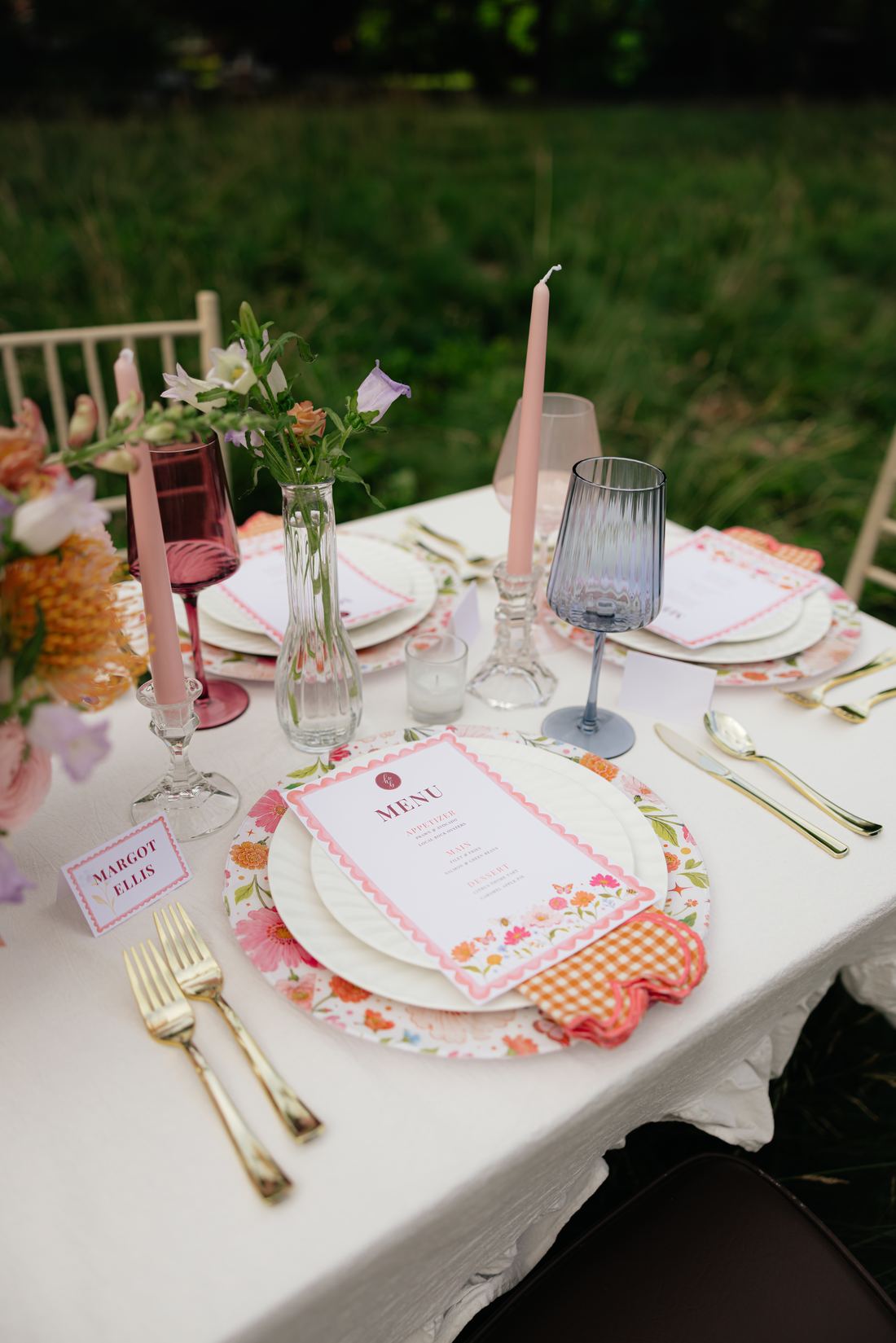 A white linen table with a green charger, coral florals and place cards at dusk