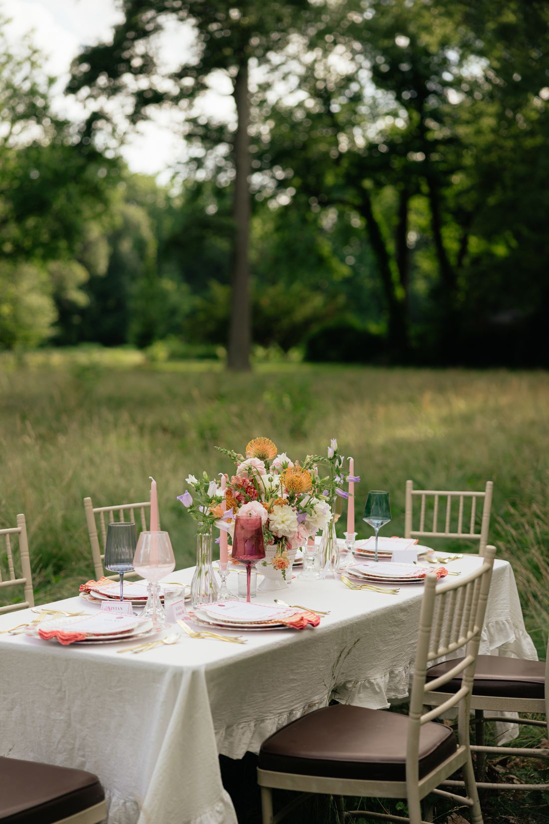 A white-linen table and gold chairs set with peach florals in a mown field